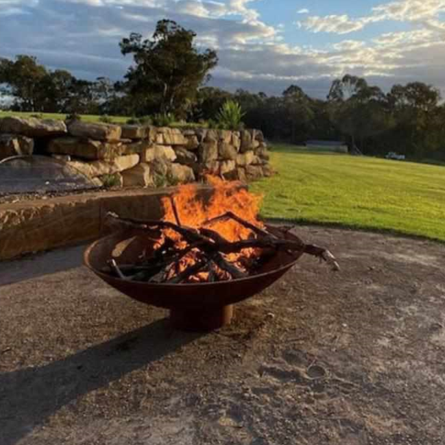 The Cauldron Fire Pit 1500mm burning a fire and half grill in background