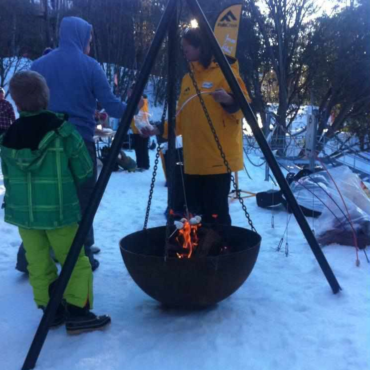 The Tripod Fire Pit in the snow with people close by toasting marshmellows